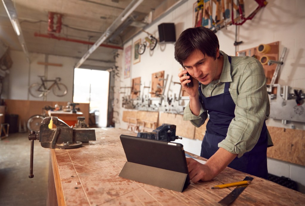 A man working in a woodshop answers a phone and works on a laptop